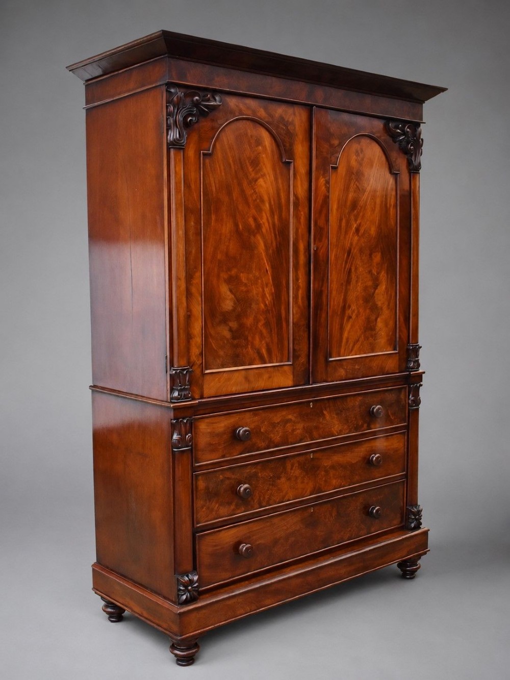 A William IV flame mahogany linen press with a carved scroll cresting, double arched panel doors, and a lower chest of drawers on turned feet. View III