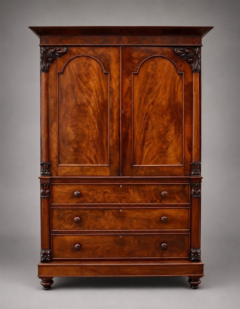 A William IV flame mahogany linen press with a carved scroll cresting, double arched panel doors, and a lower chest of drawers on turned feet. View IV