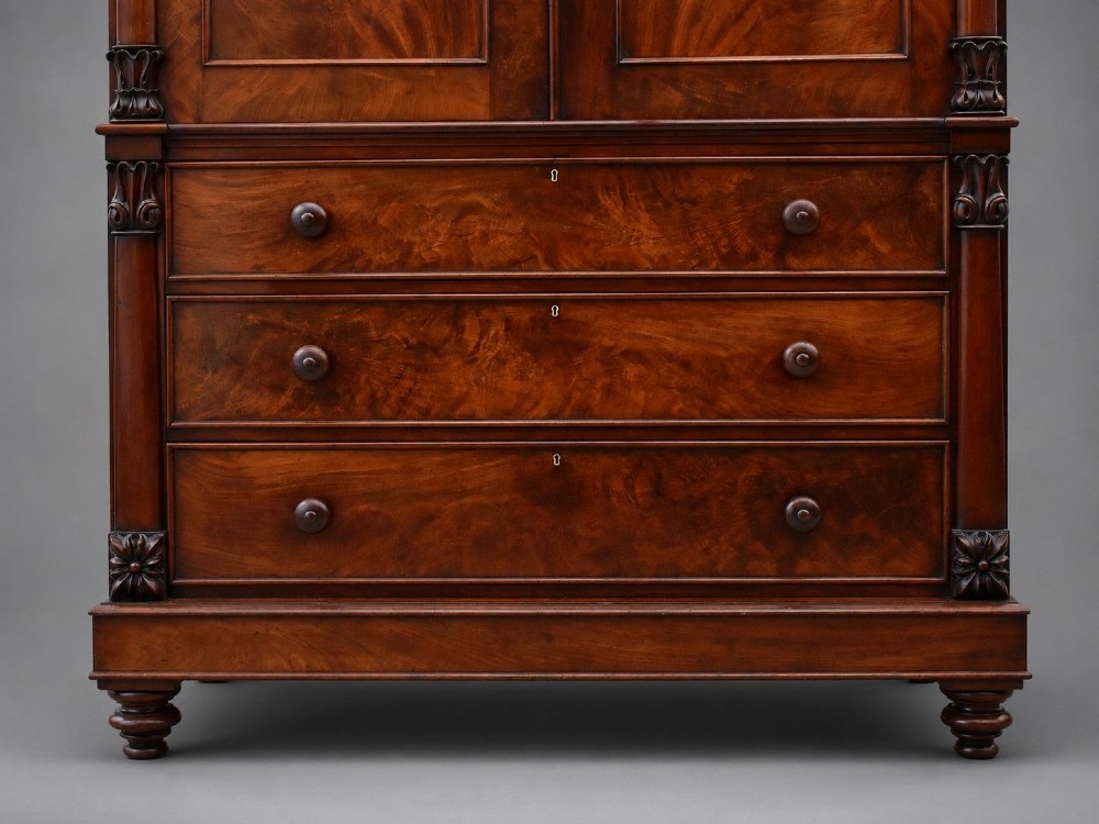A William IV flame mahogany linen press with a carved scroll cresting, double arched panel doors, and a lower chest of drawers on turned feet. View VI