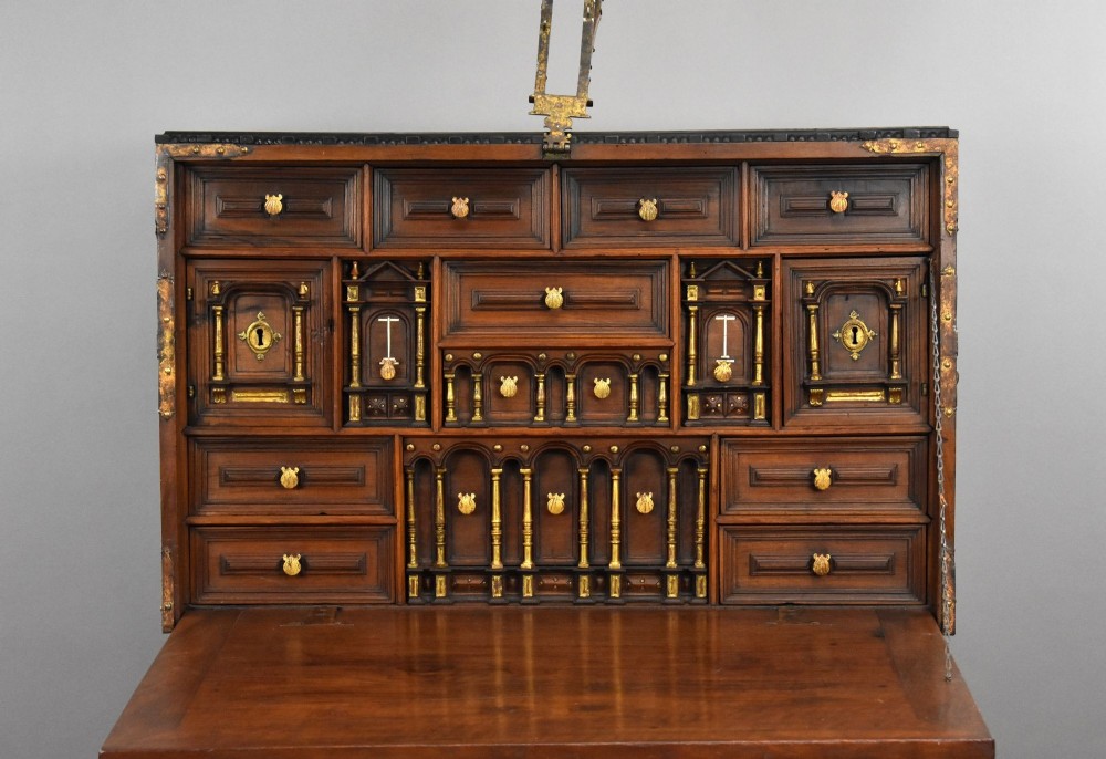 A 17th-century Spanish walnut Bargueno cabinet featuring an intricate brass lockplate and side mounts, resting on an ornate barley-twist wooden stand. View X