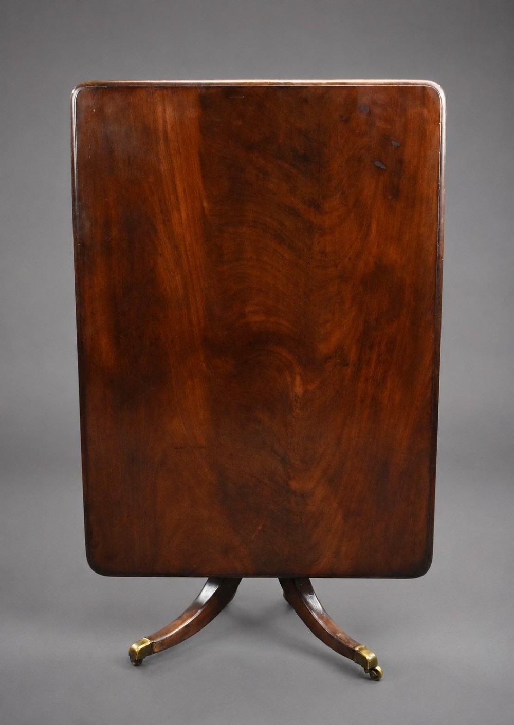 A Regency mahogany tilt-top breakfast table with its rectangular top secured in a vertical position above a pedestal base with curved splayed legs and brass castors.