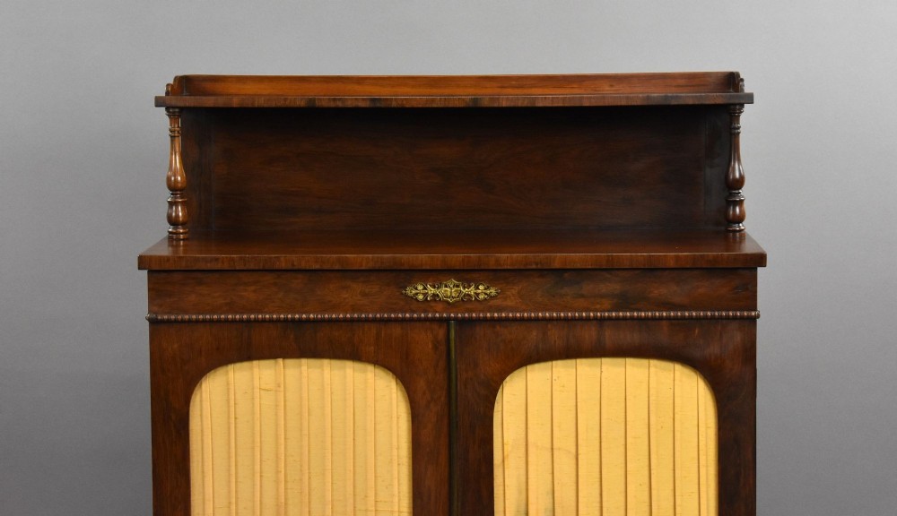 A Victorian dark rosewood chiffonier featuring an upper display shelf supported by turned pillars above lower cabinet doors fitted with pleated fabric panels. View V