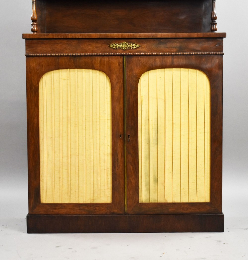 A Victorian dark rosewood chiffonier featuring an upper display shelf supported by turned pillars above lower cabinet doors fitted with pleated fabric panels. View VI