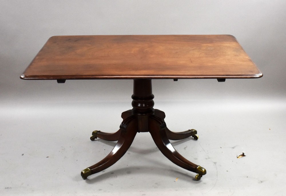 A Regency mahogany tilt-top breakfast table with its rectangular top secured in a vertical position above a pedestal base with curved splayed legs and brass castors. View V