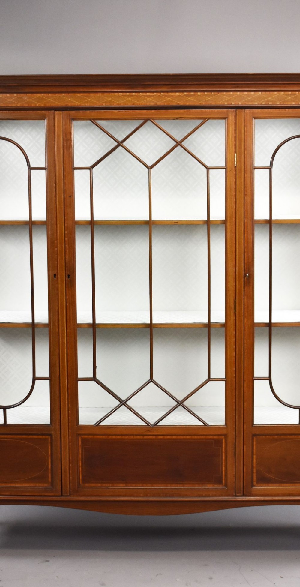 An Edwardian mahogany display cabinet featuring three glazed doors with geometric astragal glazing bars, satinwood inlay panels, and tapered legs. View III