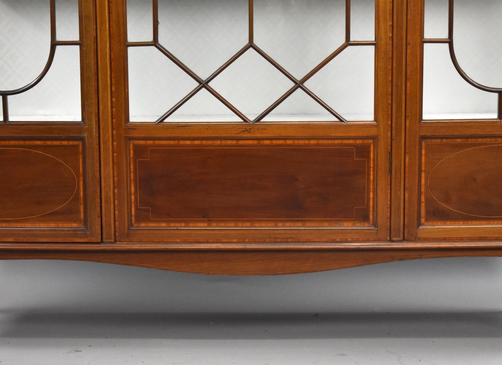 An Edwardian mahogany display cabinet featuring three glazed doors with geometric astragal glazing bars, satinwood inlay panels, and tapered legs. View VI