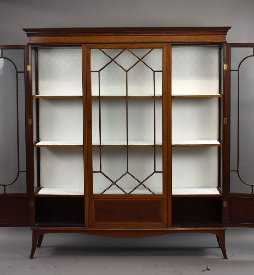 An Edwardian mahogany display cabinet featuring three glazed doors with geometric astragal glazing bars, satinwood inlay panels, and tapered legs. View IX