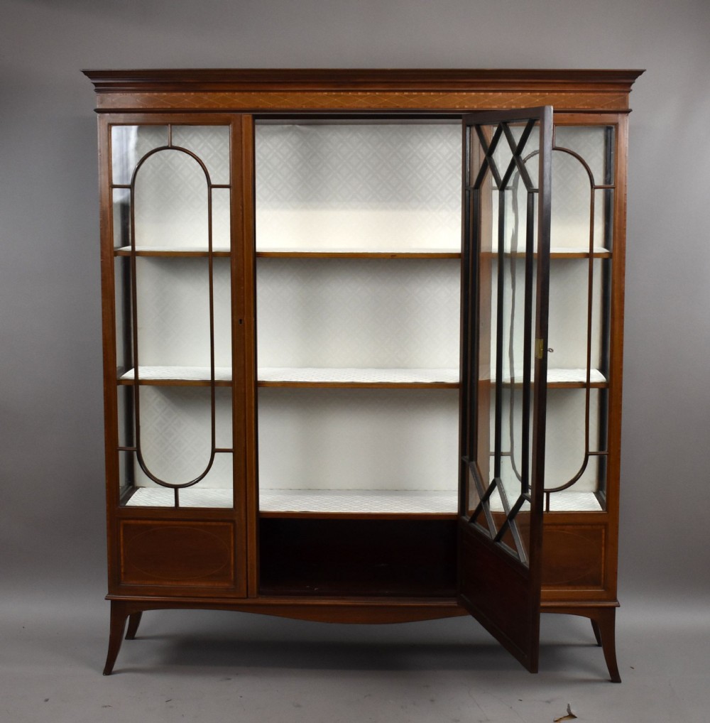An Edwardian mahogany display cabinet featuring three glazed doors with geometric astragal glazing bars, satinwood inlay panels, and tapered legs. View X