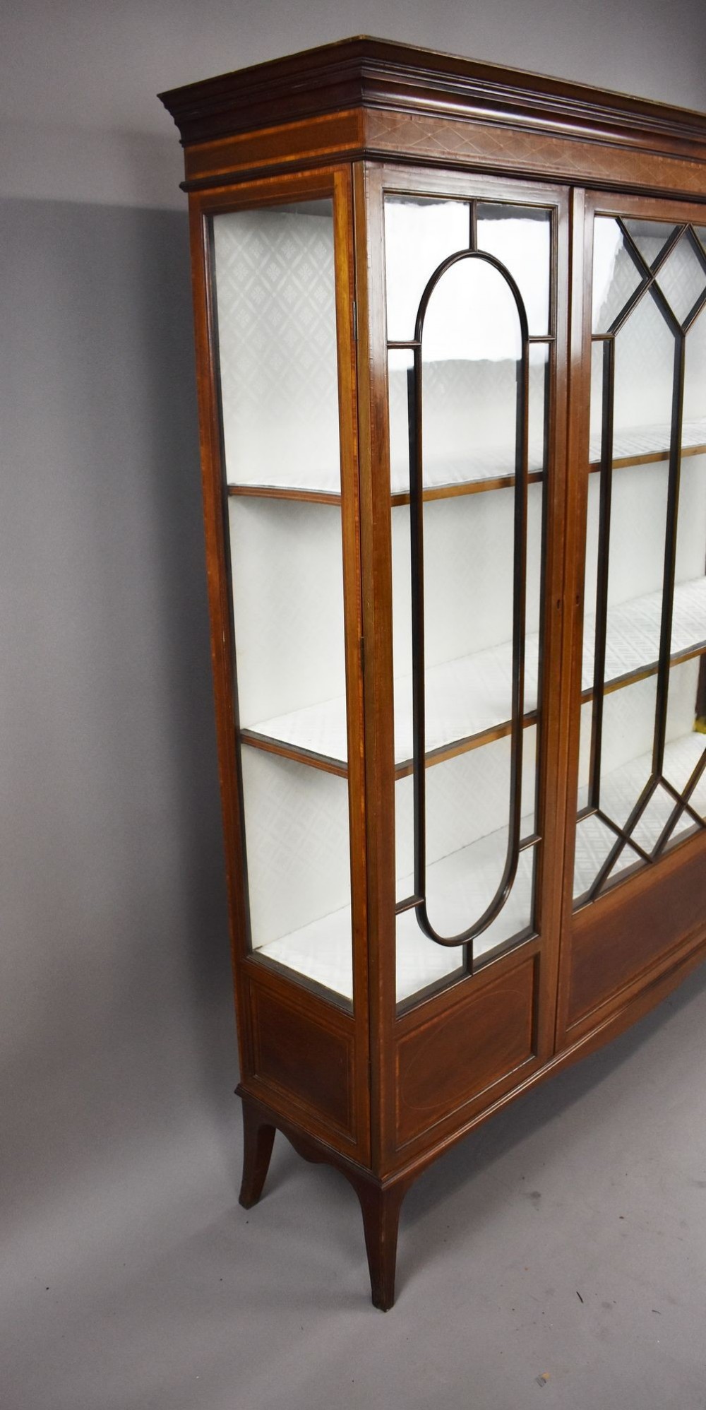 An Edwardian mahogany display cabinet featuring three glazed doors with geometric astragal glazing bars, satinwood inlay panels, and tapered legs. View XI