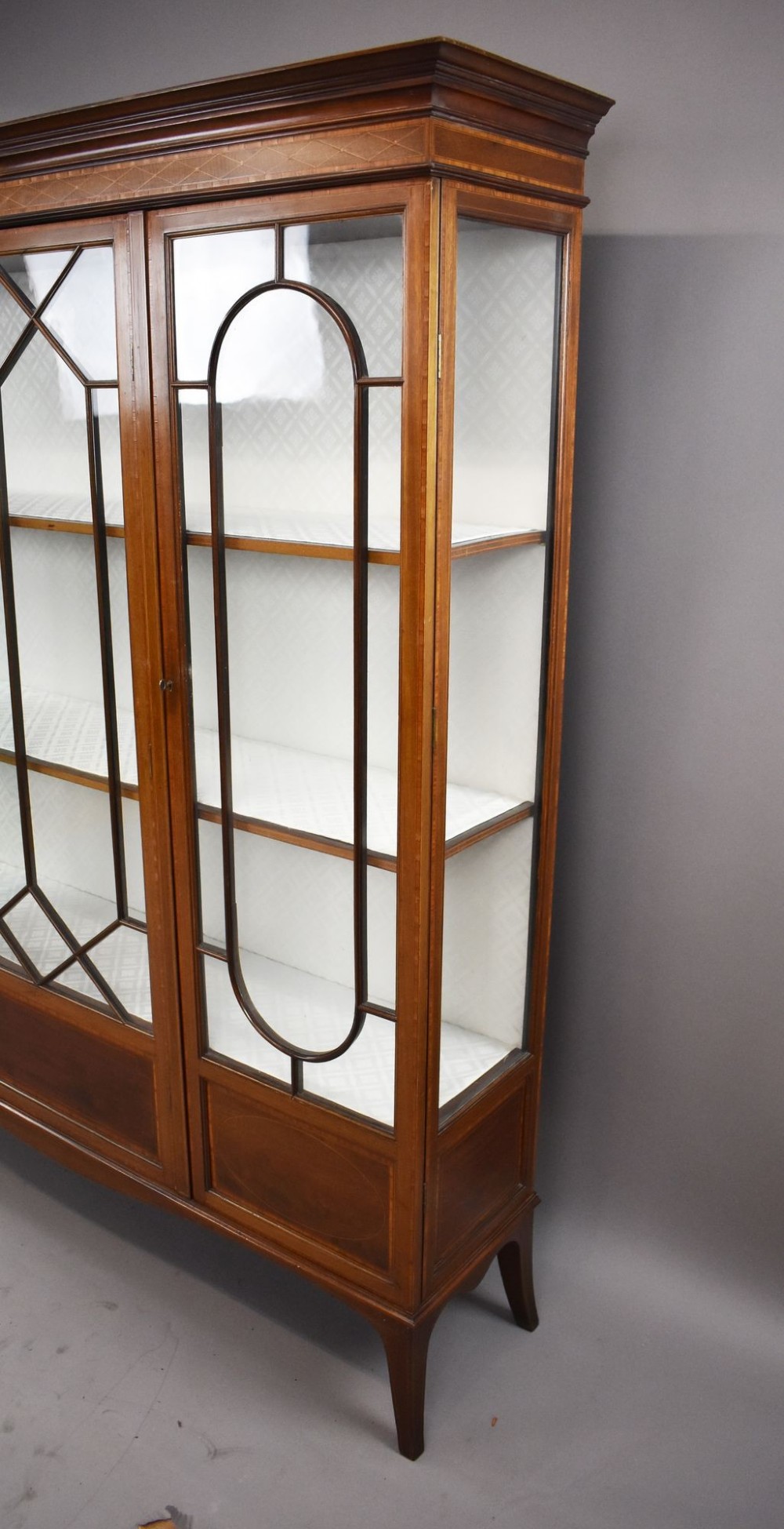 An Edwardian mahogany display cabinet featuring three glazed doors with geometric astragal glazing bars, satinwood inlay panels, and tapered legs. View XII