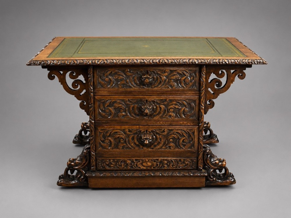 A dark oak Victorian writing desk featuring a green leather inset top with gold tooling, detailed lion mask carvings on the drawer fronts, and scroll corbels.