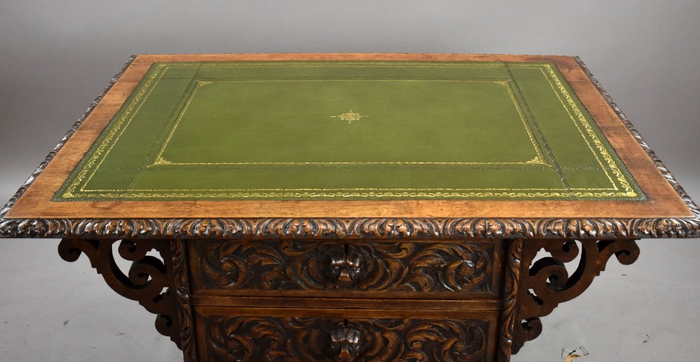 A dark oak Victorian writing desk featuring a green leather inset top with gold tooling, detailed lion mask carvings on the drawer fronts, and scroll corbels. View IV