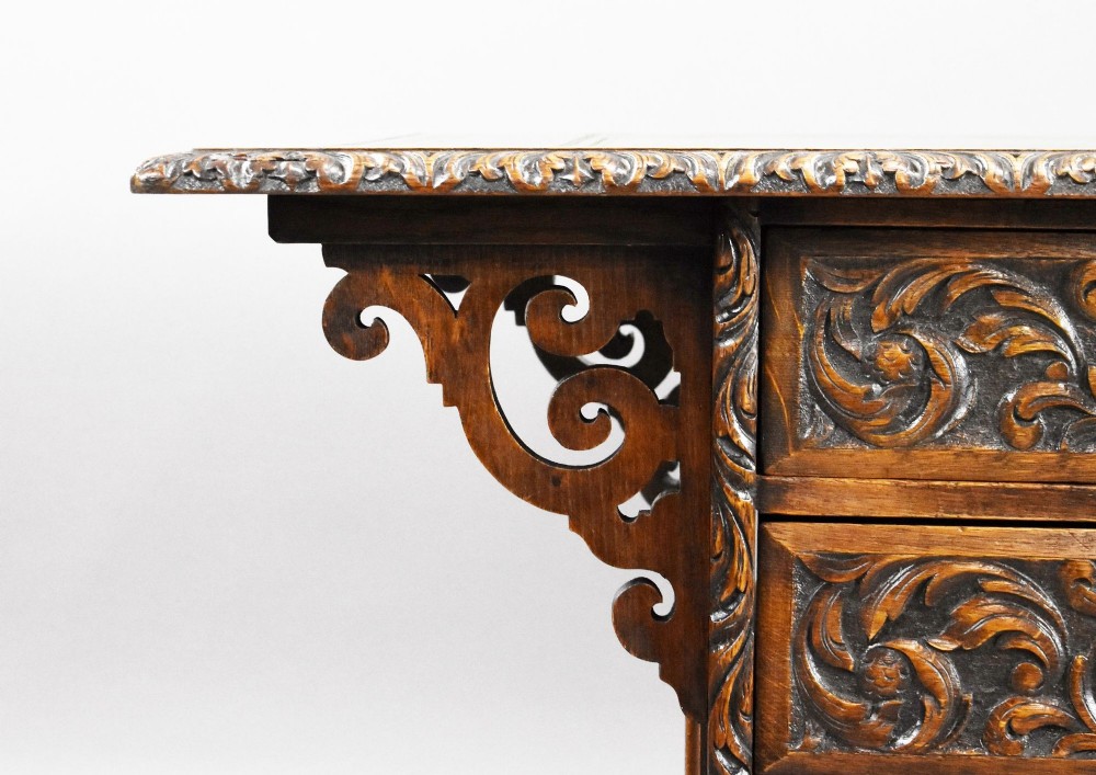 A dark oak Victorian writing desk featuring a green leather inset top with gold tooling, detailed lion mask carvings on the drawer fronts, and scroll corbels. View V