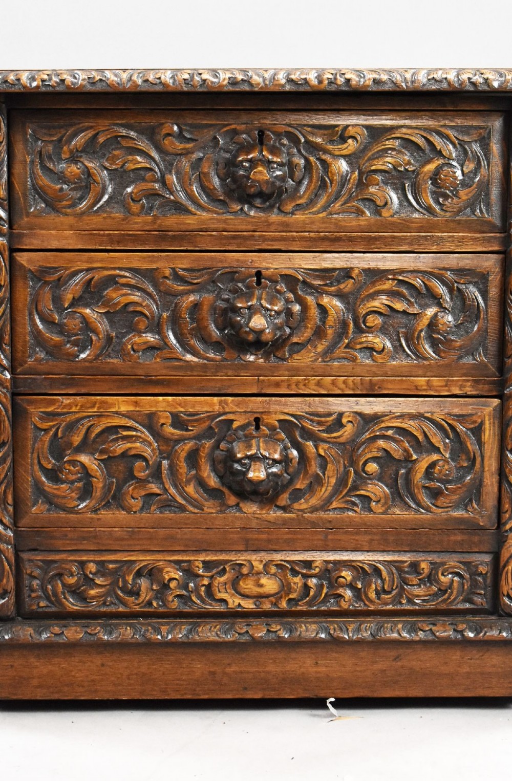 A dark oak Victorian writing desk featuring a green leather inset top with gold tooling, detailed lion mask carvings on the drawer fronts, and scroll corbels. View VII