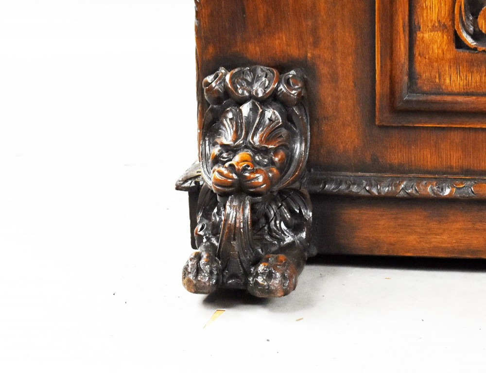 A dark oak Victorian writing desk featuring a green leather inset top with gold tooling, detailed lion mask carvings on the drawer fronts, and scroll corbels. View IX