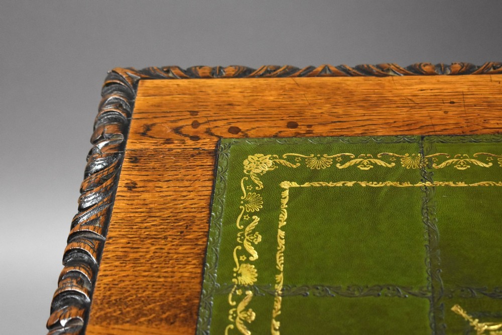 A dark oak Victorian writing desk featuring a green leather inset top with gold tooling, detailed lion mask carvings on the drawer fronts, and scroll corbels. View XI