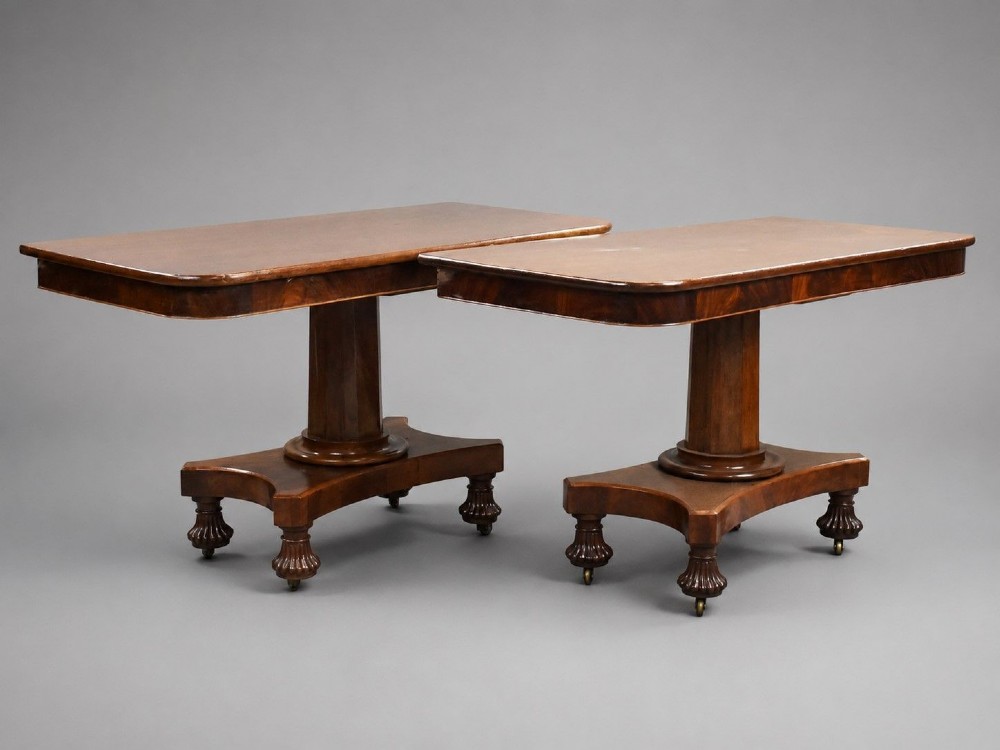 A pair of antique William IV mahogany console tables featuring rounded rectangular tops, central cylindrical pedestals, concave plinth bases, and carved lobed feet on brass castors.