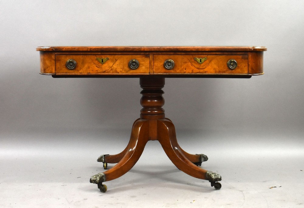 A Victorian burr walnut writing table featuring a green leather inset top, frieze drawers, and a central turned pedestal on splayed legs with brass lion paw castors. View II