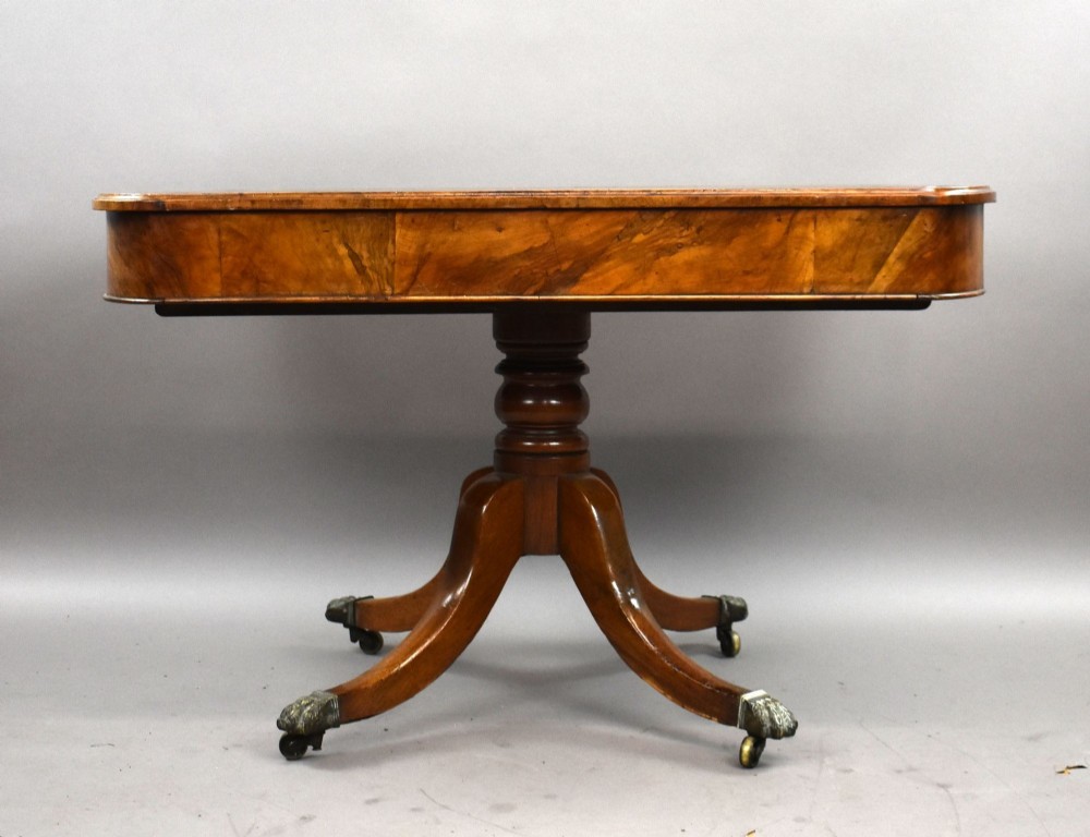 A Victorian burr walnut writing table featuring a green leather inset top, frieze drawers, and a central turned pedestal on splayed legs with brass lion paw castors. View IV