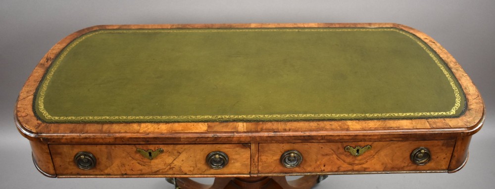 A Victorian burr walnut writing table featuring a green leather inset top, frieze drawers, and a central turned pedestal on splayed legs with brass lion paw castors. View VI