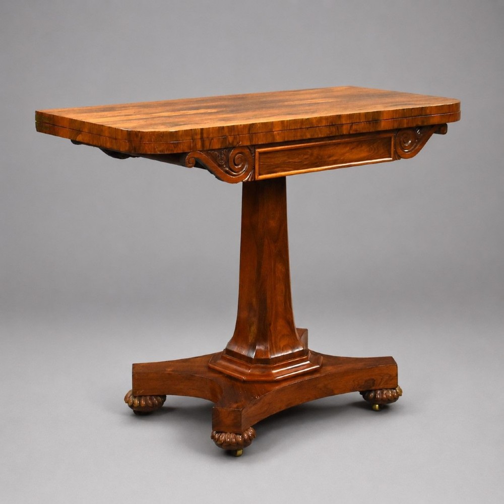A William IV rosewood fold-over card table featuring a polished rectangular top, scroll-carved frieze brackets, a faceted central pedestal, and a shaped quadform base with carved feet on castors.