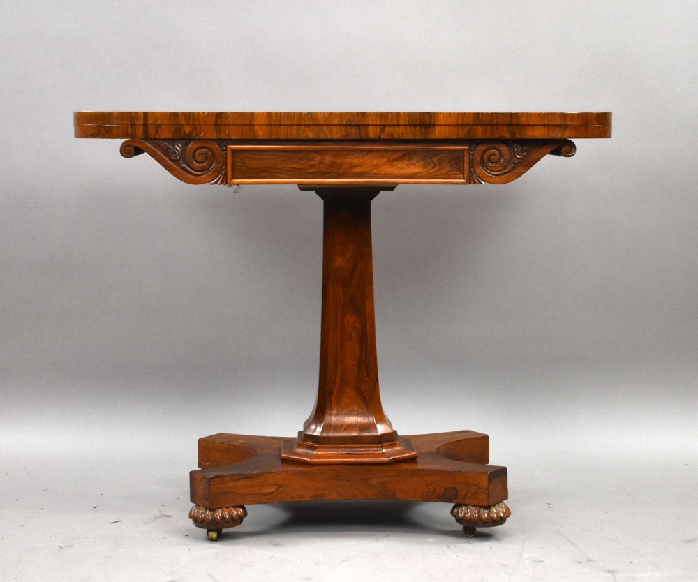 A William IV rosewood fold-over card table featuring a polished rectangular top, scroll-carved frieze brackets, a faceted central pedestal, and a shaped quadform base with carved feet on castors. View II