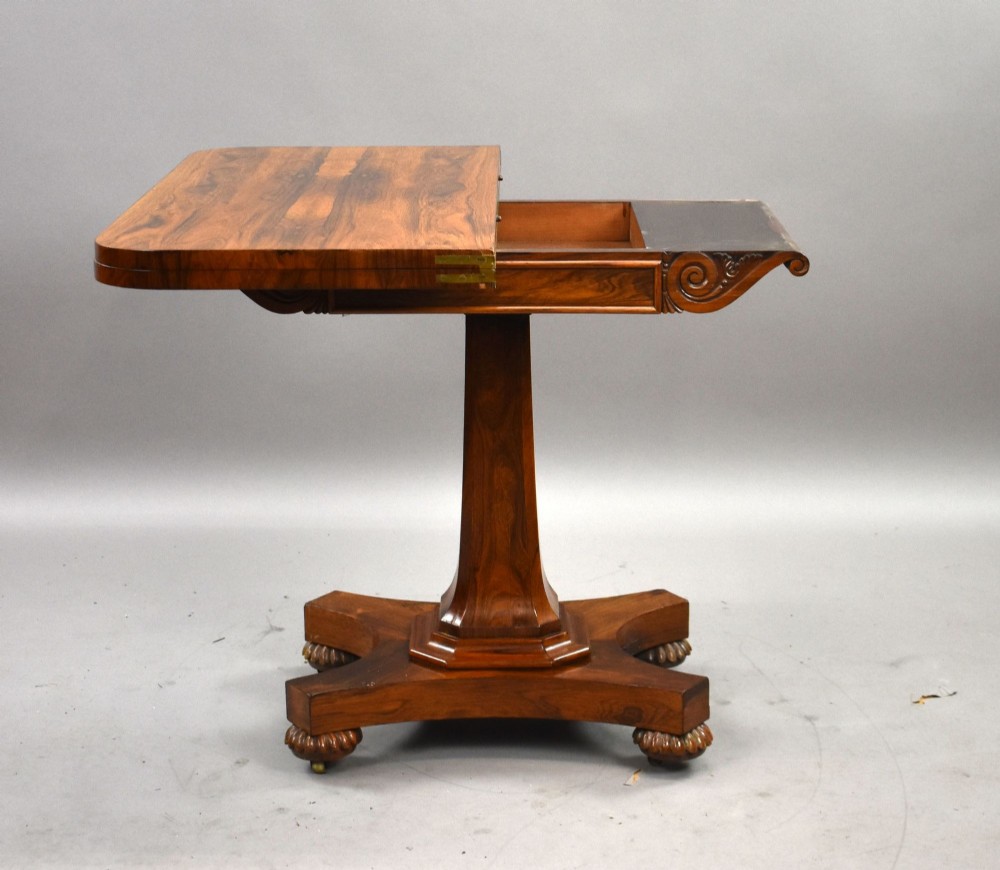 A William IV rosewood fold-over card table featuring a polished rectangular top, scroll-carved frieze brackets, a faceted central pedestal, and a shaped quadform base with carved feet on castors. View III