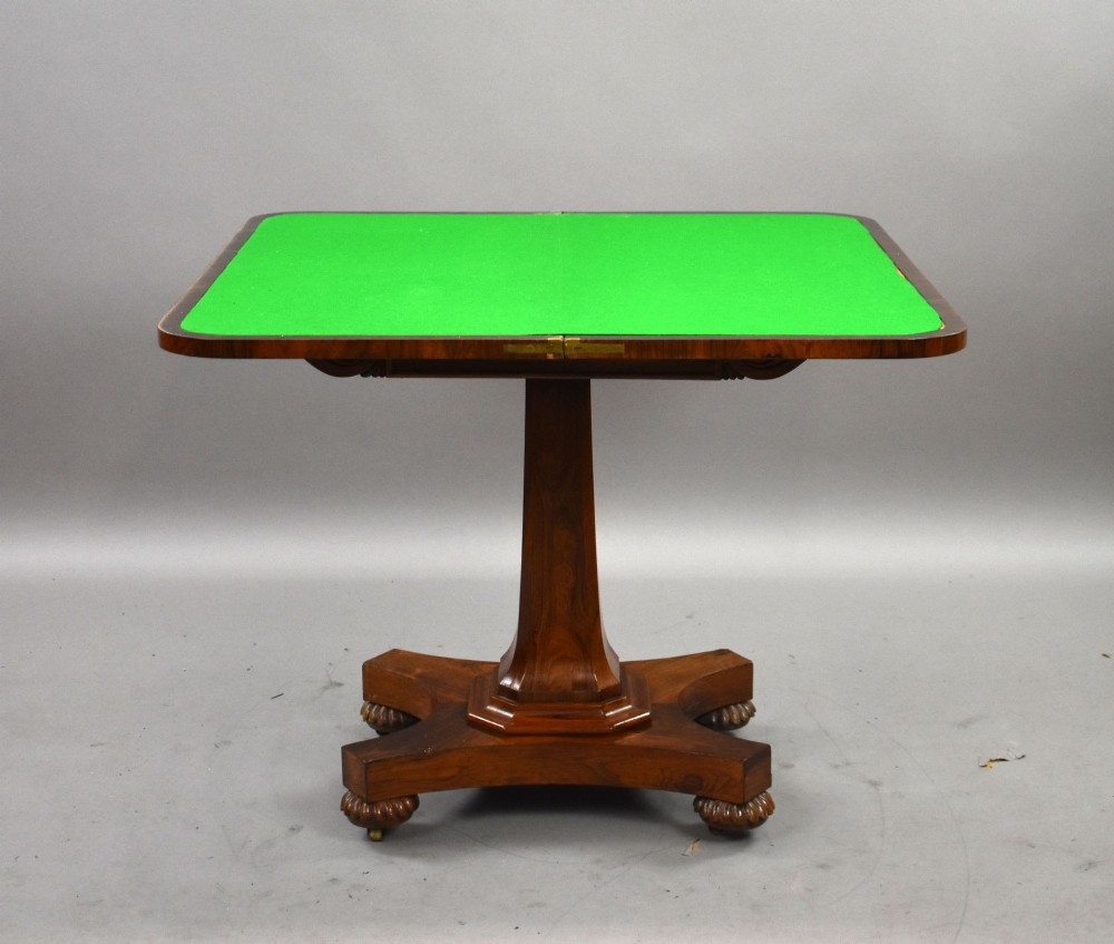 A William IV rosewood fold-over card table featuring a polished rectangular top, scroll-carved frieze brackets, a faceted central pedestal, and a shaped quadform base with carved feet on castors. View IV