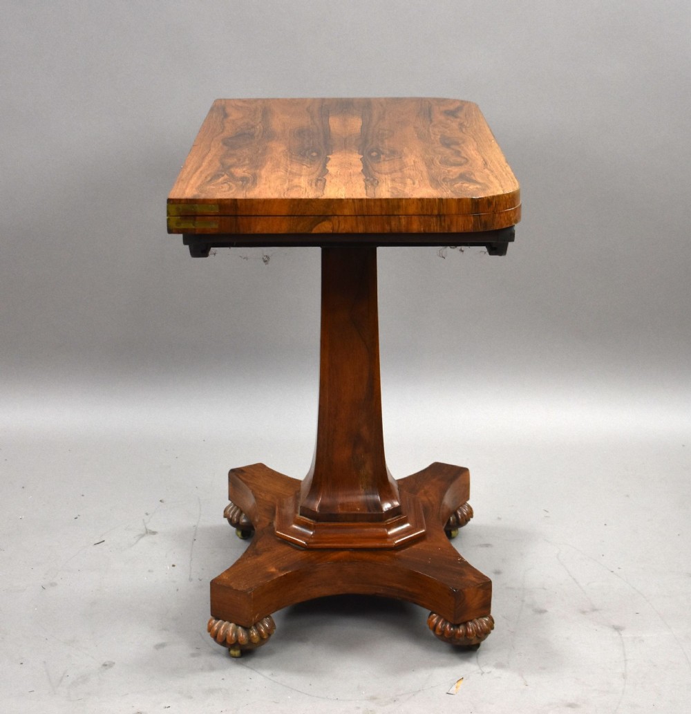 A William IV rosewood fold-over card table featuring a polished rectangular top, scroll-carved frieze brackets, a faceted central pedestal, and a shaped quadform base with carved feet on castors. View VI