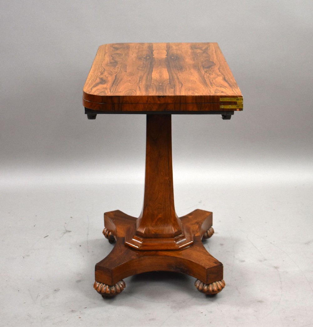 A William IV rosewood fold-over card table featuring a polished rectangular top, scroll-carved frieze brackets, a faceted central pedestal, and a shaped quadform base with carved feet on castors. View VII