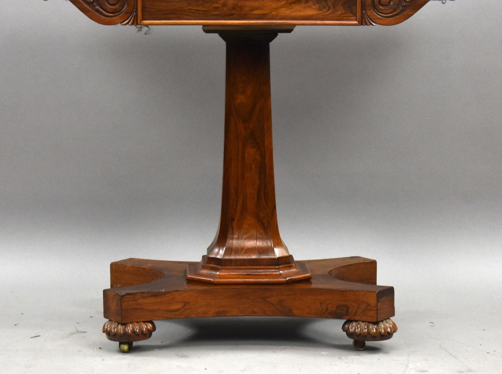 A William IV rosewood fold-over card table featuring a polished rectangular top, scroll-carved frieze brackets, a faceted central pedestal, and a shaped quadform base with carved feet on castors. View VIII