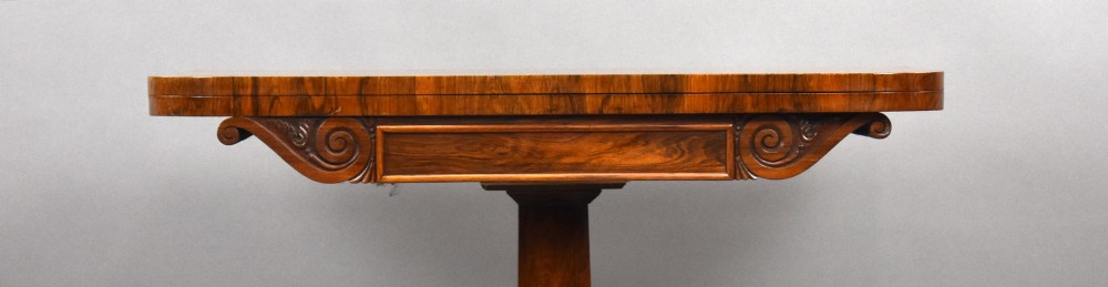 A William IV rosewood fold-over card table featuring a polished rectangular top, scroll-carved frieze brackets, a faceted central pedestal, and a shaped quadform base with carved feet on castors. View IX