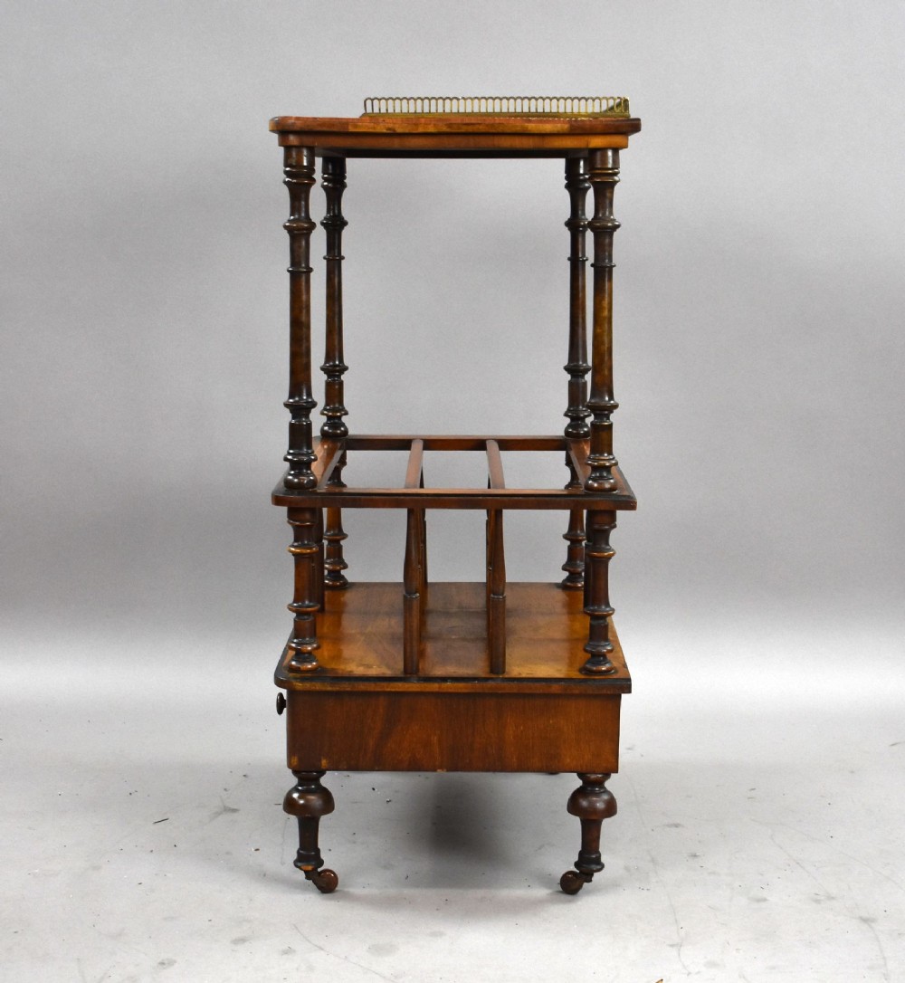 A Victorian burr walnut Canterbury featuring a top tier with a brass gallery rail, turned spindle columns, open partitions, a single inlaid drawer, and turned legs on brass castors. View IV