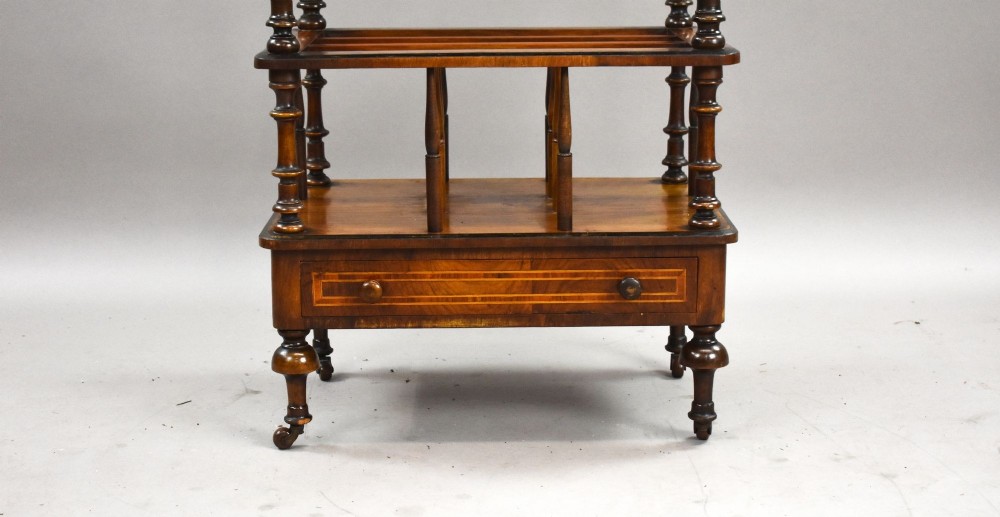 A Victorian burr walnut Canterbury featuring a top tier with a brass gallery rail, turned spindle columns, open partitions, a single inlaid drawer, and turned legs on brass castors. View VI