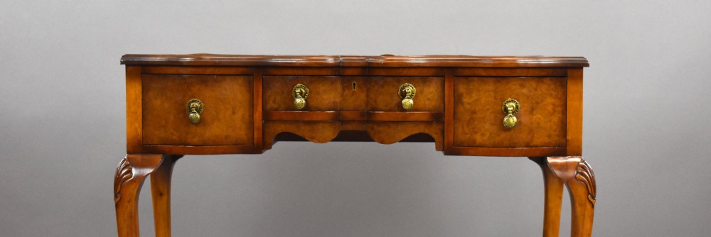A Queen Anne style walnut writing desk featuring a brown leather inset top, serpentine front drawers with brass handles, and carved cabriole legs. View VI