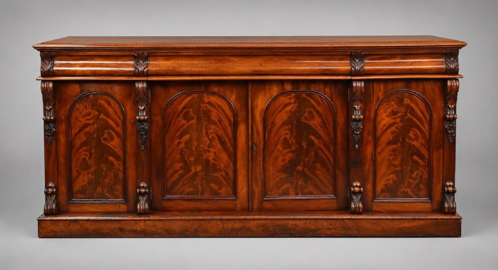 A nineteenth-century Victorian flame mahogany four-door sideboard featuring arched door panels, carved pilaster corbels, and a wide plinth base.