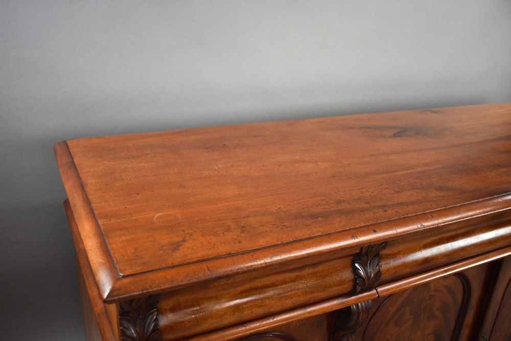 A nineteenth-century Victorian flame mahogany four-door sideboard featuring arched door panels, carved pilaster corbels, and a wide plinth base. View V