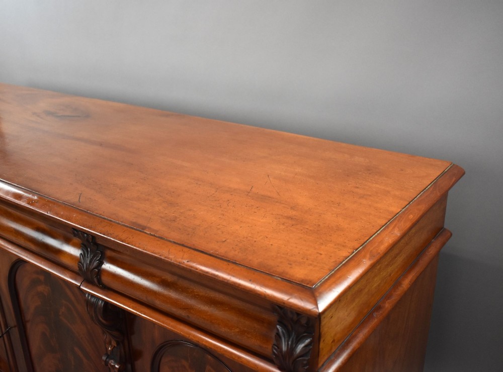 A nineteenth-century Victorian flame mahogany four-door sideboard featuring arched door panels, carved pilaster corbels, and a wide plinth base. View VII