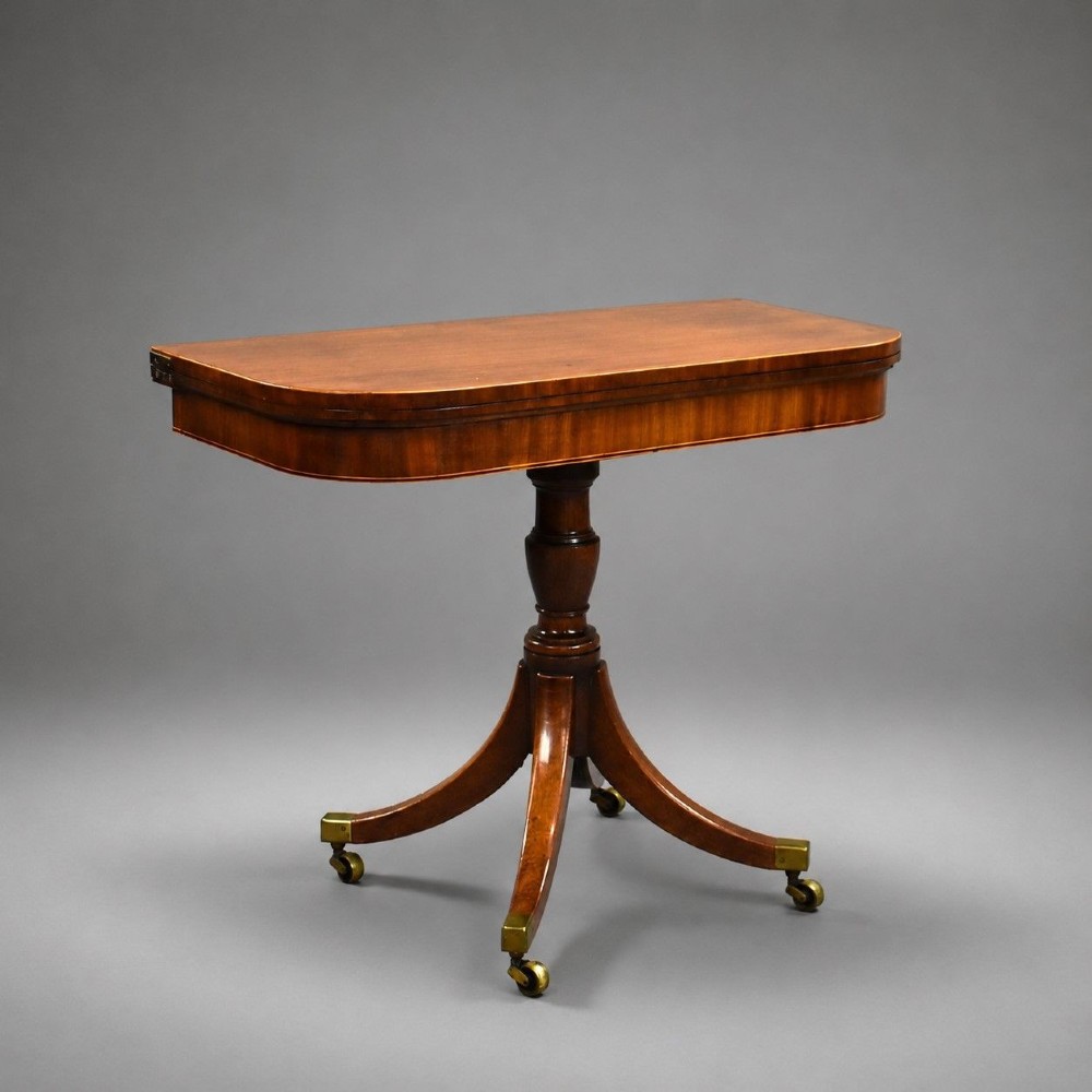 A Regency mahogany card table featuring a folding swivel top, a turned central pedestal column, and four splayed sabre legs with brass castors.