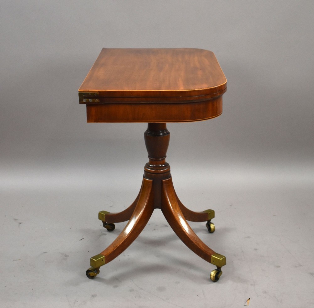 A Regency mahogany card table featuring a folding swivel top, a turned central pedestal column, and four splayed sabre legs with brass castors. View V
