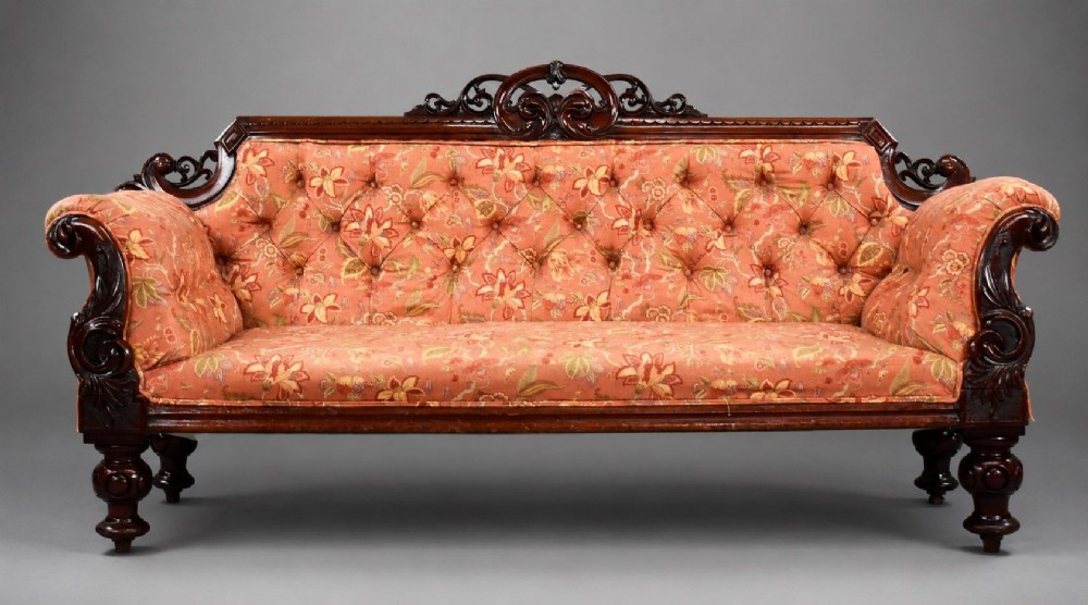 Victorian mahogany settee featuring carved scroll cresting along the top rail, button-tufted floral fabric upholstery, rolled armrests, and turned wooden front legs.