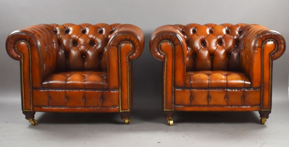 A matching pair of warm brown leather Chesterfield armchairs featuring deep button tufting, brass studding, rolled arms, and brass castors. View II
