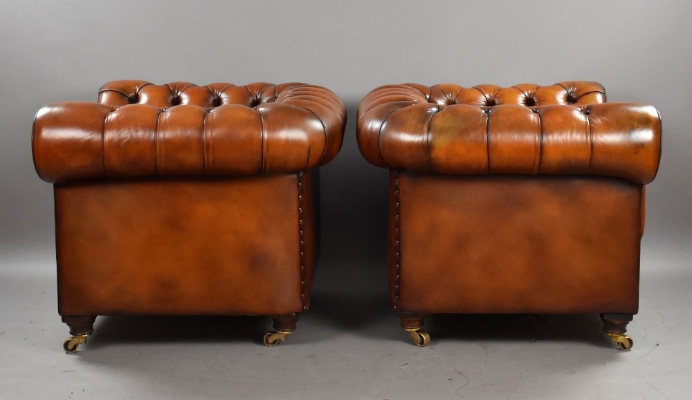 A matching pair of warm brown leather Chesterfield armchairs featuring deep button tufting, brass studding, rolled arms, and brass castors. View III