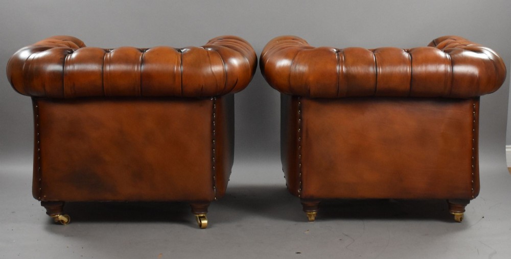A matching pair of warm brown leather Chesterfield armchairs featuring deep button tufting, brass studding, rolled arms, and brass castors. View IV