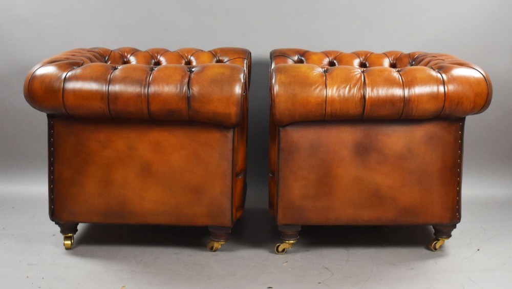 A matching pair of warm brown leather Chesterfield armchairs featuring deep button tufting, brass studding, rolled arms, and brass castors. View V