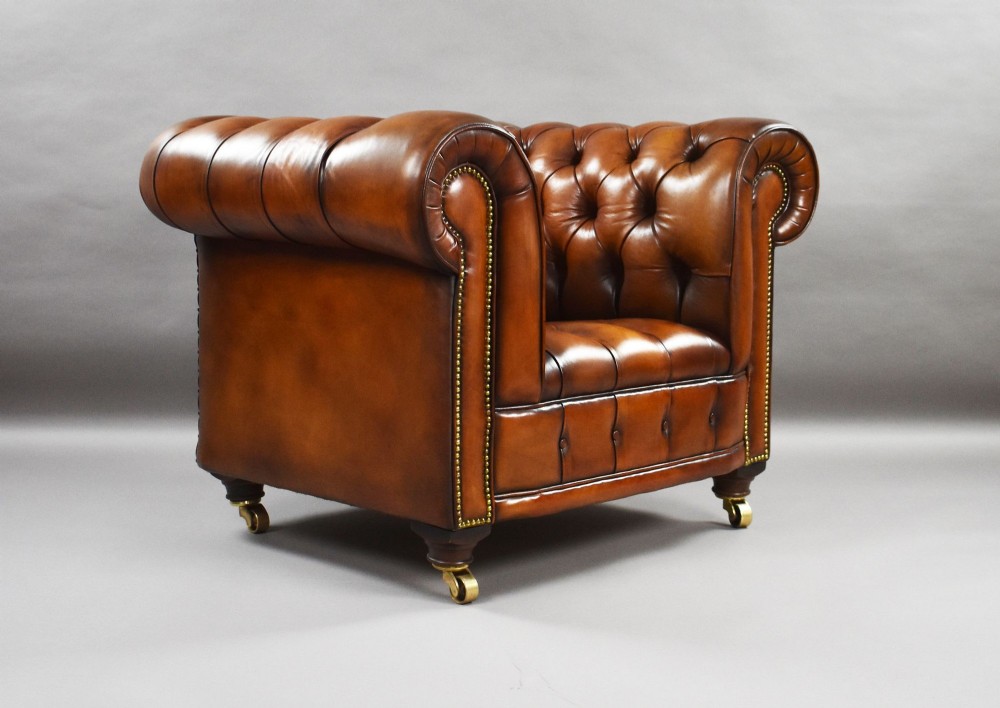 A matching pair of warm brown leather Chesterfield armchairs featuring deep button tufting, brass studding, rolled arms, and brass castors. View VI