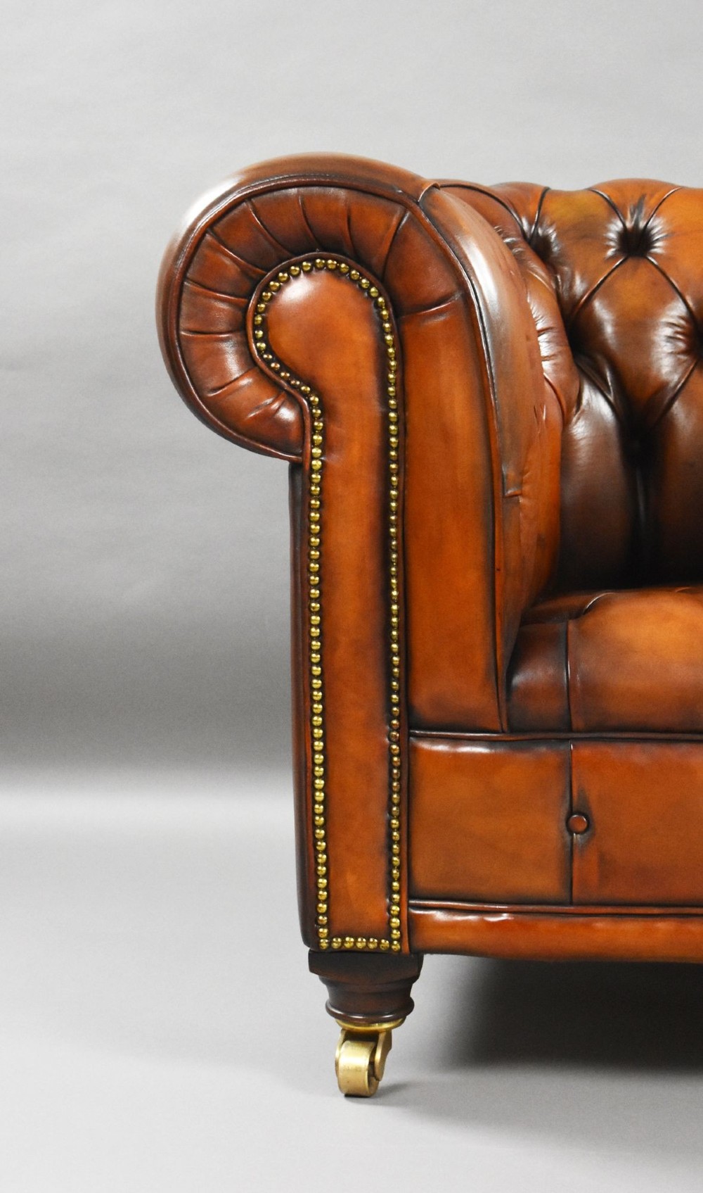 A matching pair of warm brown leather Chesterfield armchairs featuring deep button tufting, brass studding, rolled arms, and brass castors. View VIII