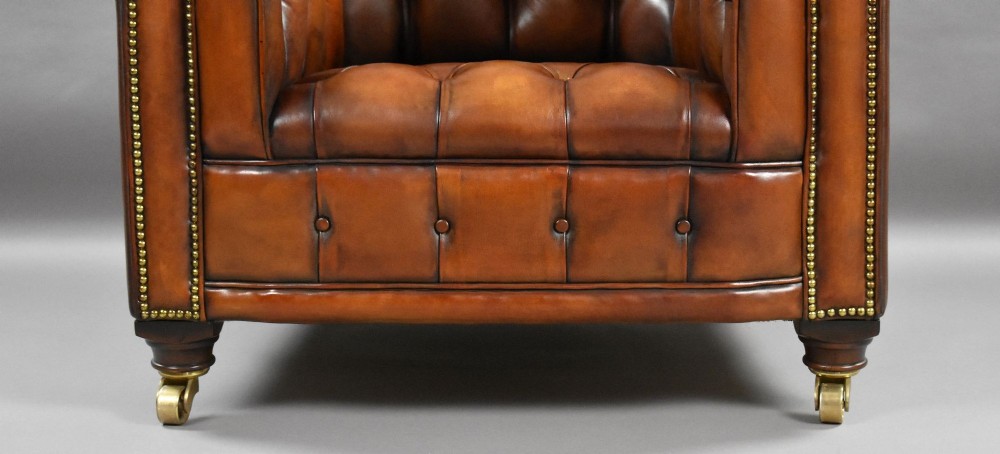 A matching pair of warm brown leather Chesterfield armchairs featuring deep button tufting, brass studding, rolled arms, and brass castors. View IX