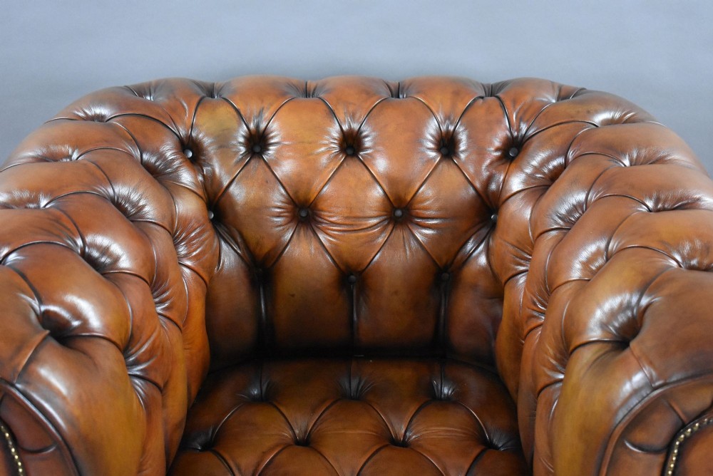 A matching pair of warm brown leather Chesterfield armchairs featuring deep button tufting, brass studding, rolled arms, and brass castors. View X