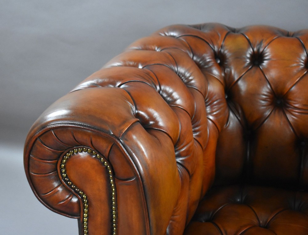 A matching pair of warm brown leather Chesterfield armchairs featuring deep button tufting, brass studding, rolled arms, and brass castors. View XI